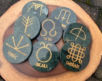 May include: Eight green marble coasters with gold painted symbols and text. The symbols represent the eight sabbats of the Wheel of the Year. The text on the coasters reads: Imbolc, Ostara, Litha, Lughnasadh, Mabon, Samhain, Yule, and  Alban Arthan.