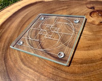 May include: A clear glass coaster with a geometric design featuring circles and lines. The coaster has rounded feet and is set on a wooden surface. The design is a white, intricate pattern.