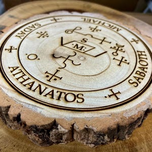 May include: A circular wooden coaster with a laser-etched design. The design features text and symbols in a circular arrangement. The coaster has a natural wood grain edge and is light brown in colour.