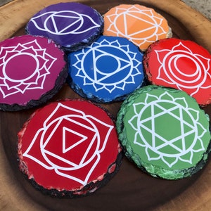 Op de afbeelding: Zeven ronde, leisteen onderzetters met witte chakra symbolen. De onderzetters zijn geschilderd in de kleuren van de zeven chakra's: rood, oranje, geel, groen, blauw, indigo en violet.