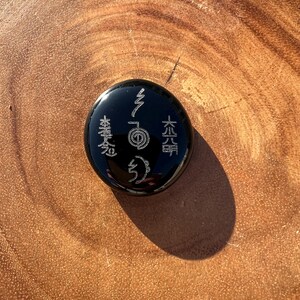 以下が含まれることがあります： 刻まれたシンボルと文字が施された、丸い黒い石。石は滑らかで反射する表面を持っています。シンボルは明るい色で、暗い背景と対照的です。石は木製の表面に置かれています。