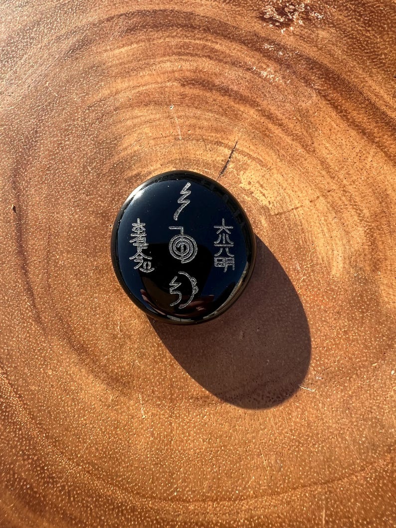 以下が含まれることがあります： 刻まれたシンボルと文字が施された、丸い黒い石。石は滑らかで反射する表面を持っています。シンボルは明るい色で、暗い背景と対照的です。石は木製の表面に置かれています。