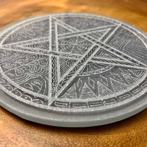 Pode incluir: Um descanso de copo redondo de vidro com um pentagrama branco gravado na superfície. O pentagrama é rodeado por uma variedade de desenhos intrincados, incluindo espirais, linhas e formas geométricas.