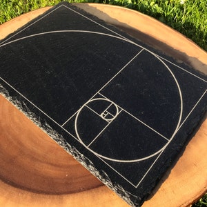 Puede incluir: Una bandeja de servir de pizarra negra con un diseño grabado en blanco de la espiral de la proporción áurea. La bandeja tiene un borde áspero y natural.