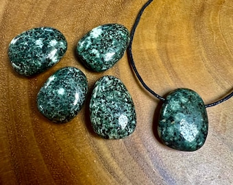 May include: Five green and white speckled stones, four loose and one strung on a black cord as a pendant. The stones are oval-shaped and polished, with a natural, organic appearance. The background is a warm-toned wooden surface.