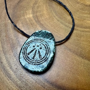 May include: A green stone pendant with a black cord. The pendant has a circular design with three lines and three dots. The stone has a mottled appearance and is set against a wooden background.