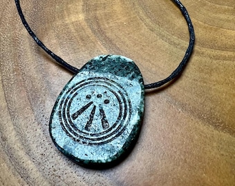 May include: A green stone pendant on a black cord. The pendant features a circular design with three lines and three dots. The stone has a mottled appearance. The pendant is set against a wooden background.