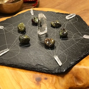 Op de afbeelding: Een zwart leisteenbord met een geometrisch patroon en zeven kristallen. De kristallen zijn in een cirkel rond een helder kwartskristal in het midden gerangschikt. De kristallen zijn groen, rood en helder.