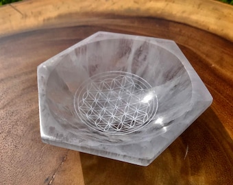 May include: A hexagonal bowl carved from clear, translucent quartz crystal. The interior base of the bowl features a finely etched white geometric pattern consisting of multiple overlapping circles arranged in a symmetrical flower of life design.