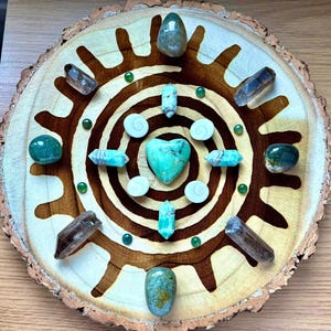 Op de afbeelding: Een ronde houten dienblad met een spiraalvormig ontwerp erop geschilderd. Het dienblad is versierd met verschillende groene en bruine kristallen, waaronder een hartvormig kristal in het midden.