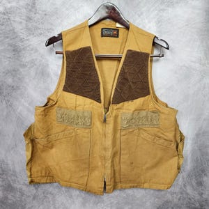 May include: A tan utility vest featuring dark brown quilted shoulder patches, a central zipper closure, and two large front pockets with textured flaps. The interior label displays the text Sears Sports Center. The vest is sleeveless.