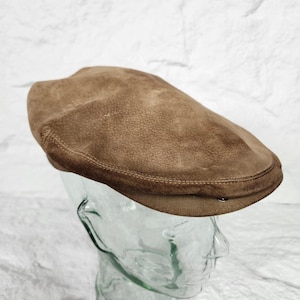 May include: A brown suede flat cap with a rounded crown and a short, stiffened brim. The cap features a soft, textured surface with visible stitching along the edge of the brim and a structured panel construction across the top.