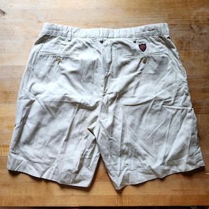 May include: Beige shorts featuring a waistband with belt loops, two back pockets with button closures, and a small red, white, and blue shield-shaped patch logo located above the right back pocket. The shorts have a visible button on each pocket.