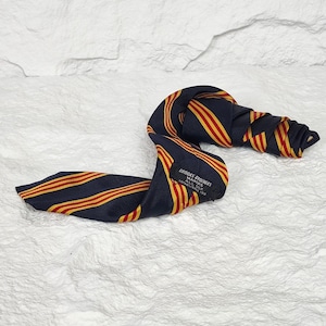 May include: A navy blue necktie featuring diagonal red and yellow stripes. The tie includes a black label stitched on the back that reads Brooks Brothers Makers All Silk Established 1818. The fabric has a visible ribbed texture.