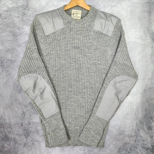 May include: A grey ribbed knit sweater featuring a crew neckline, long sleeves, and tonal fabric patches on the shoulders and elbows. The interior collar tag displays the text "MADE IN ENGLAND" and other details. The garment has a textured finish.