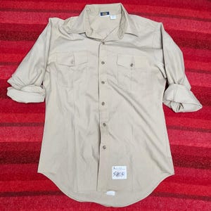 May include: A long-sleeved tan button-down shirt featuring a classic collar, two chest pockets with buttoned flaps, and a central button placket. The sleeves are rolled up, and a small white rectangular tag with handwritten text is attached near the hem.