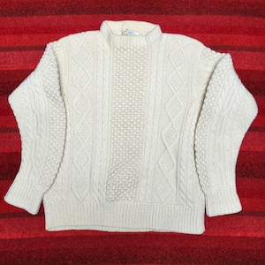 May include: An off-white long-sleeve sweater featuring a complex knit pattern. The front displays a central vertical panel of honeycomb texture flanked by vertical cable knit columns, while the sleeves showcase a dense, textured knit design.