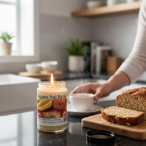 May include: A clear glass jar candle with a lit wick and a label reading Banana Nut Bread, positioned next to a wooden cutting board holding a loaf of sliced banana bread and a white ceramic teacup on a saucer on a dark countertop.