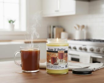 May include: A clear glass jar candle with a yellow wax base and a label reading Rum & Butter, positioned next to a steaming glass mug of dark brown liquid. The candle features a lit wick and a black metal lid resting on a white cloth napkin.