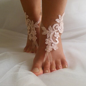 May include: A pair of delicate, white lace barefoot sandals with a floral design. The sandals are tied with a ribbon around the ankle and have a delicate, feminine look.