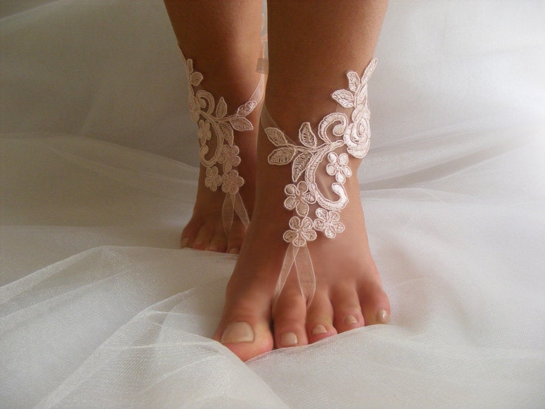 May include: A pair of delicate, white lace barefoot sandals with a floral design. The sandals are tied with a ribbon around the ankle and have a delicate, feminine look.