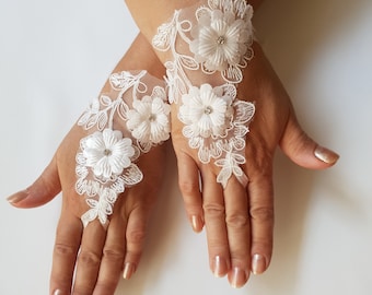 May include: A pair of white lace bridal gloves with delicate floral appliques and small pearl-like embellishments. The gloves are fingerless and extend to the wrist.