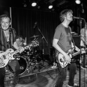 May include: A black and white photo of a band performing on stage. The lead singer is on the right, holding a black electric guitar and singing into a microphone. The guitarist on the left is playing a black electric guitar. The drummer is in the background.