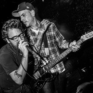 May include: Two people, one with a microphone and one playing a black electric guitar with a white pickguard. The guitar has the text "Fender Telecaster" on the headstock.