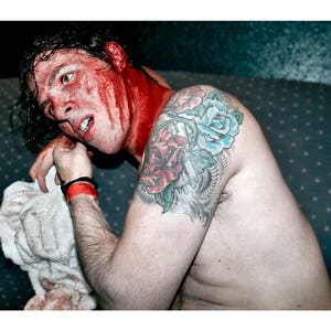 May include: A man with a bloody face and a floral tattoo on his shoulder. The tattoo features red, blue, and green flowers.