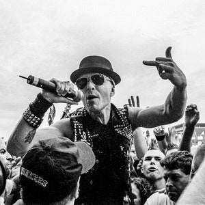 May include: A black and white photo of a musician performing on stage. The musician is wearing a black hat, sunglasses, and a studded leather jacket. The musician is holding a microphone and singing into it. The musician is surrounded by a crowd of people.