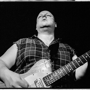 May include: A man in a plaid shirt plays a black electric guitar with a white pickguard. He is looking up and smiling.
