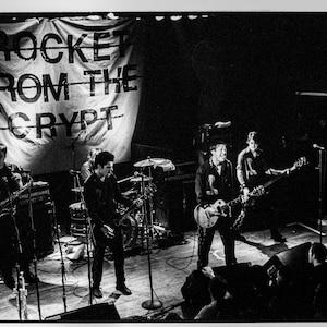 May include: Black and white photo of a band performing on stage. The band is playing in front of a banner that says "ROCKET FROM THE CRYPT".