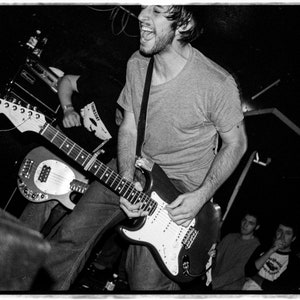May include: A black and white photo of a person playing an electric guitar on stage. The person is wearing a grey t-shirt and jeans. The guitar is a Fender Stratocaster.