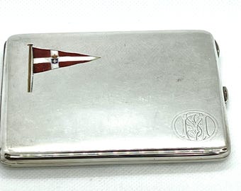 May include: A silver cigarette case with a flag design. The flag is triangular, with a white stripe, a red stripe, and a crest. The case has a rounded edge and a clasp. The case has an engraved monogram.