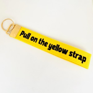May include: A bright yellow fabric keychain strap featuring the black text Pull on the yellow strap. The strap is attached to a gold-toned metal clasp and a circular split ring, providing a functional accessory for keys or bags with a bold design.