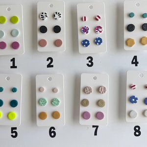 May include: A collection of eight sets of stud earrings displayed on white cards. Each card features three pairs of earrings in various colors and designs, including solid colors, patterns, and geometric shapes. The earrings are numbered 1 through 8.