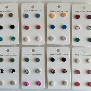 Op de afbeelding: Twaalf paar kleine, ronde, kleioorbellen die op witte kaartjes worden getoond. Elk kaartje heeft de tekst "clay + collection" erop gedrukt. De oorbellen zijn in verschillende kleuren en patronen, waaronder effen kleuren, geometrische patronen en bloemmotieven.