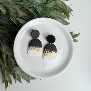 May include: A pair of black and white statement earrings with gold accents. The earrings are shaped like arches and have a white base with a black top and gold flecks.