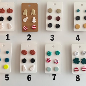 May include: Eight sets of stud earrings displayed on white and brown cards, numbered one through eight. The collection features various shapes including pumpkins, ghosts, circles, hexagons, flowers, clovers, and striped patterns in diverse colors.
