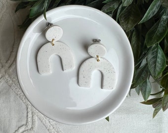 May include: A pair of white speckled arch-shaped earrings with gold accents. The earrings are on a white plate.