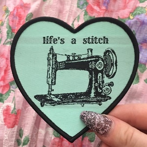 Puede incluir: Un parche en forma de corazón con borde negro y centro verde claro. El parche presenta una ilustración negra de una máquina de coser vintage y el texto "life's a stitch" en negro. El fondo es un estampado floral rosa.
