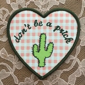 May include: Heart-shaped patch with a pink and white gingham design. It features a green cactus illustration and the phrase "don't be a prick" in black script. The patch has a dark green border.