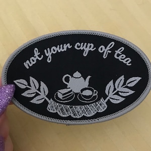 May include: Oval black patch with the phrase "not your cup of tea" in white script. The patch features a teapot, teacup, and cake design. The patch is trimmed with a gray border.