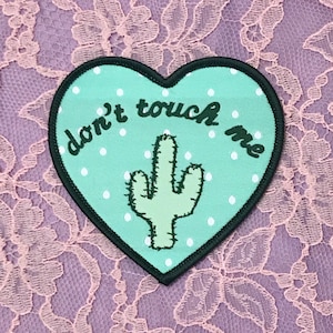 May include: A heart-shaped patch with a teal background and white polka dots. The patch features a green cactus illustration and the words "don't touch me" in black script. The patch has a black border.