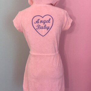 Peut inclure: Robe à capuche rose clair avec des manches courtes et une ceinture assortie. Le dos de la robe présente un cœur violet avec les mots "Angel Baby" brodés à l'intérieur. La robe est en tissu éponge doux.