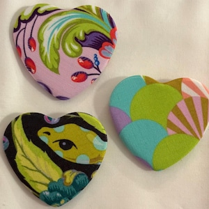 May include: Three heart-shaped fabric patches with colorful patterns. One patch has a pink background with green, purple, and red floral designs. Another features a black background with yellow and green leaf and eye designs. The third has a blue, green, and pink geometric pattern.