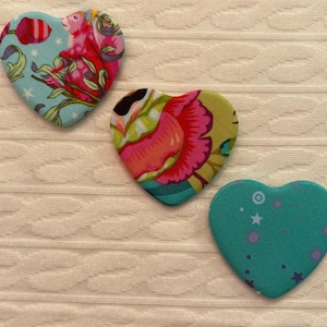 Może przedstawiać: Trzy naszywki z tkaniny w kształcie serca. Jedna jest jasnoniebieska z kwiatowym i małpim wzorem. Druga ma różowo-żółty wzór kwiatowy. Trzecia jest turkusowa z fioletowymi gwiazdami i kółkami.