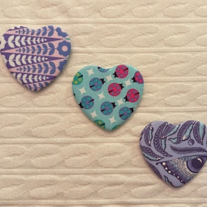 May include: Three heart-shaped fabric patches with patterned designs. One features a blue and purple floral pattern, another has a turquoise background with ladybugs, and the third showcases a purple and blue feather and eye design.