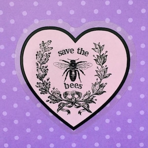 Könnte beinhalten: Ein herzförmiger Aufkleber mit dem Text "save the bees" und einer Bienenillustration. Das Design in Schwarz und Weiß ist vor einem hellrosa Hintergrund mit Lorbeerzweigen und einem Band platziert. Der Aufkleber hat einen schwarzen Rand.