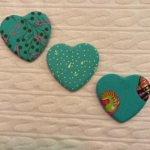 May include: Three teal heart-shaped fabric patches with unique patterns. One features pink safety pins and brown vines, another has white dots and anchors, and the third showcases a colorful design. These patches are ideal for crafting and adding a decorative touch.